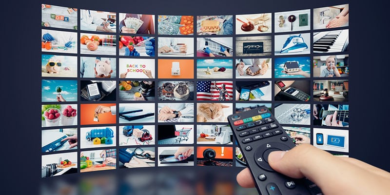 Hand holding remote flips through TV advertising on a wall of TVs.