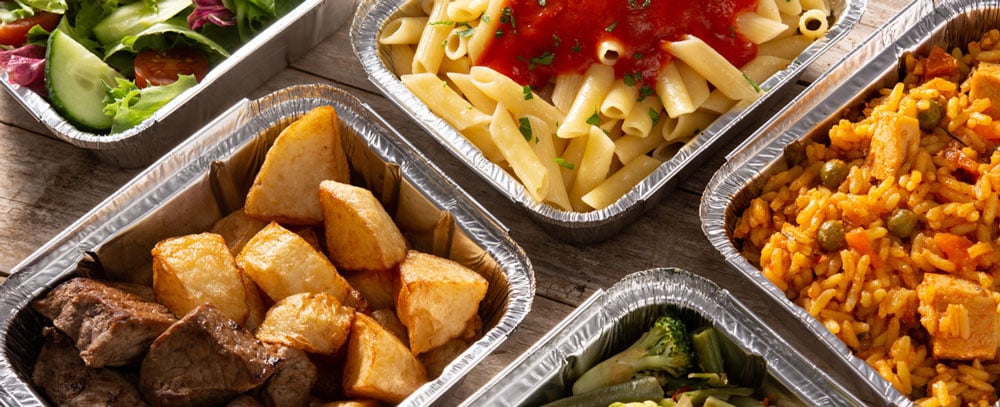 Aluminum catering containers sit on table containing pasta, potatoes, rice, beef, and more.