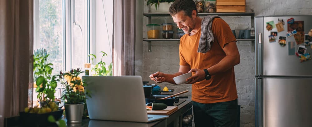 Man in orange shirt follows recipe on his laptop as he cooks a meal in his kitchen.