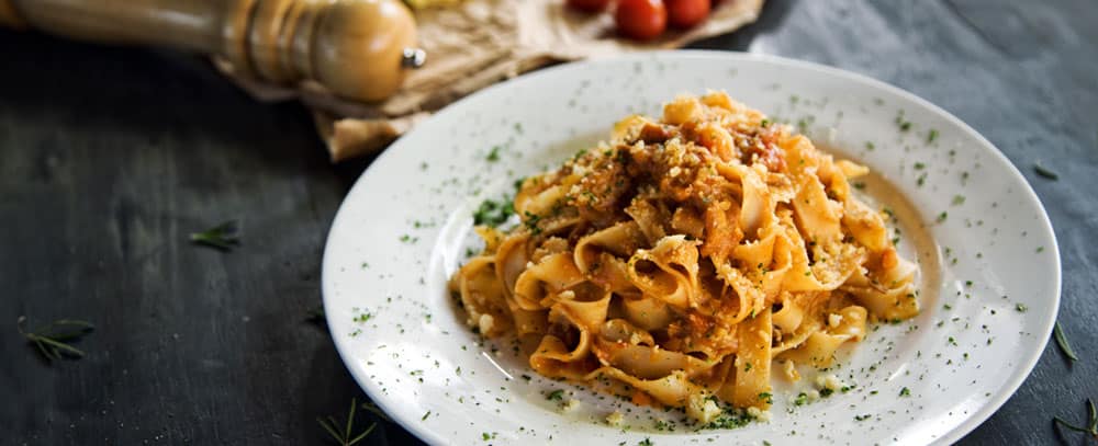 White plate of pasta carbonara is served to table with parsley decoration and pepper grinder in background.