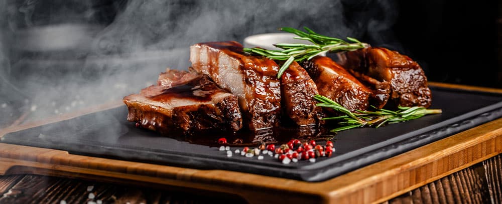 Image of a sizzling plate of glazed ribs garnished with herbs and served with a side of sauce.
