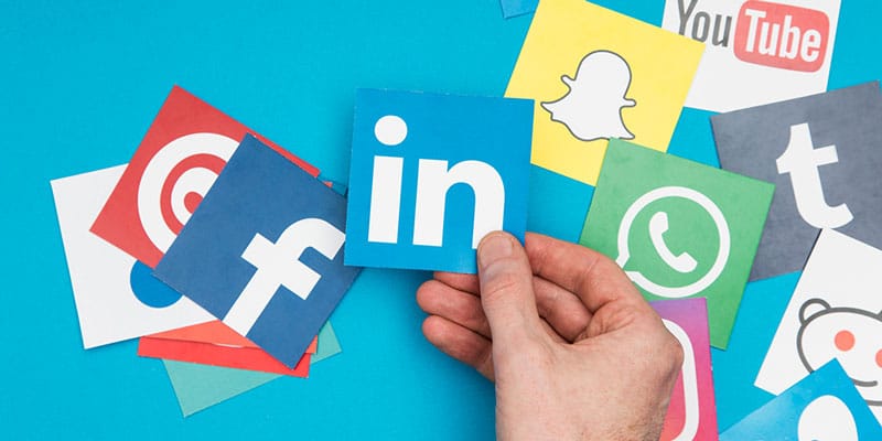 Hand adds LinkedIn to other social media platforms laid across table.